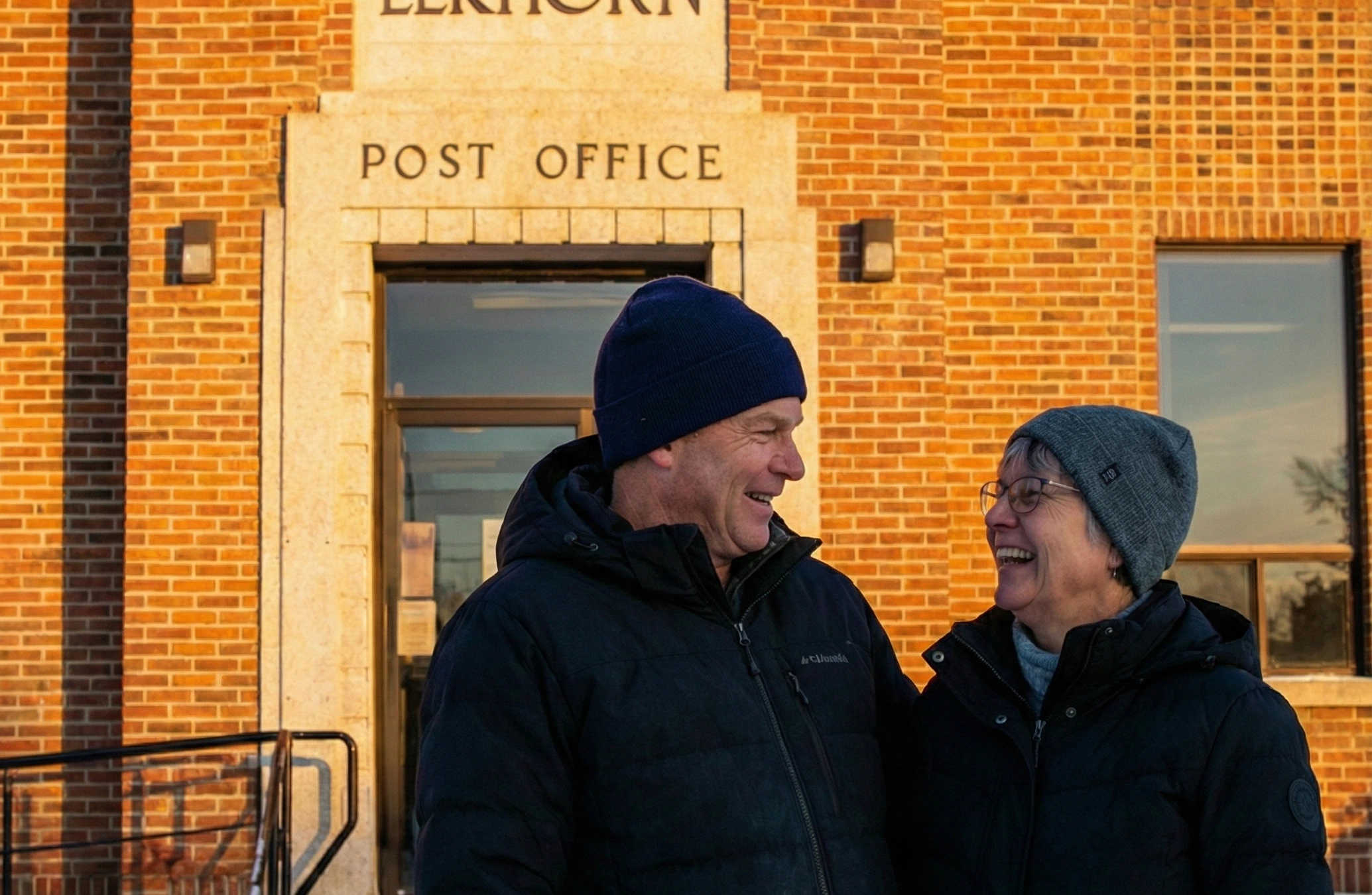 An older couple smiling and laughing together outside the Elkhorn Post Office, a brick building, on a sunny day in Elkhorn, Manitoba.