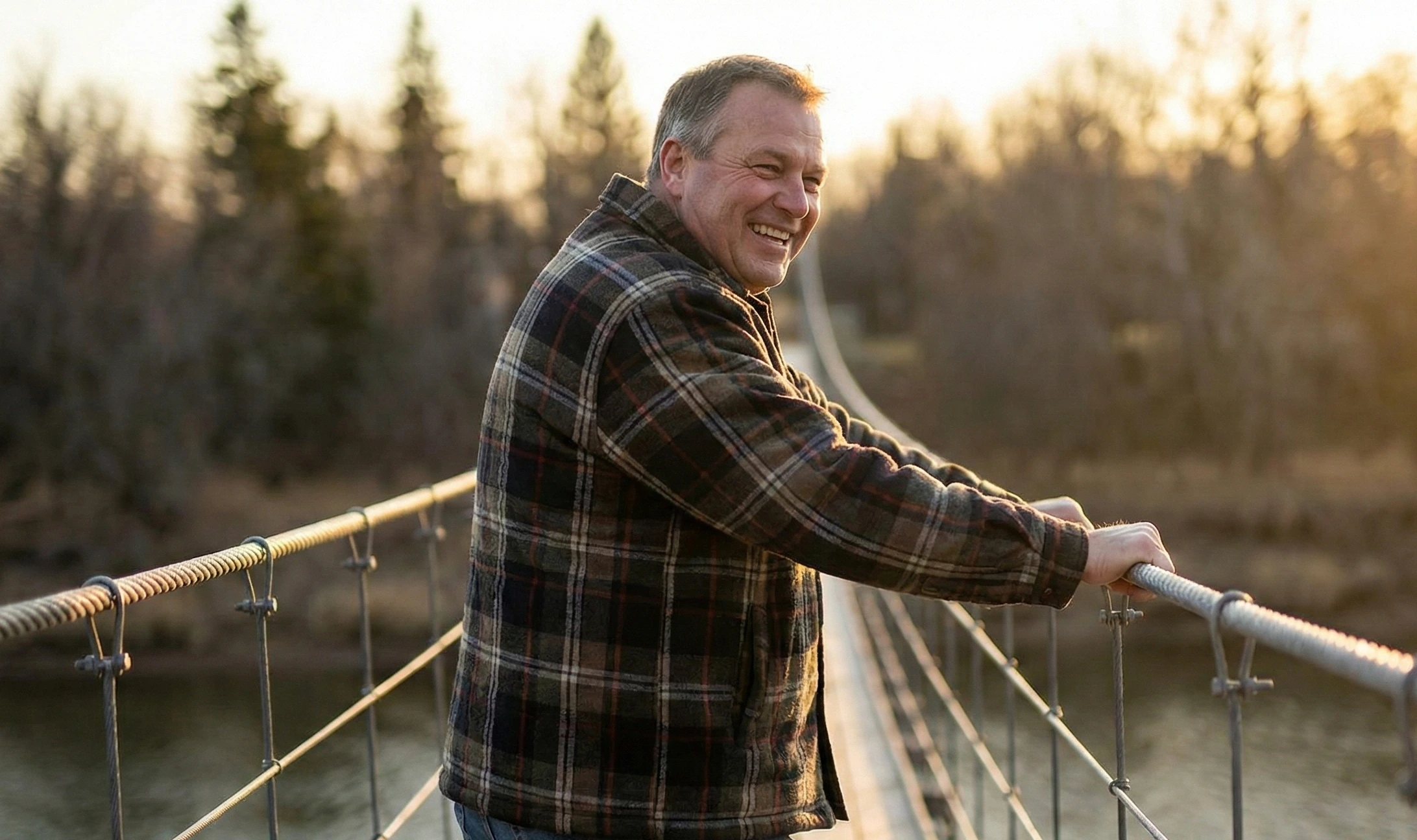 A smiling middle-aged man leaning on a railing of a suspension bridge in Souris, Manitoba, surrounded by trees in autumn.